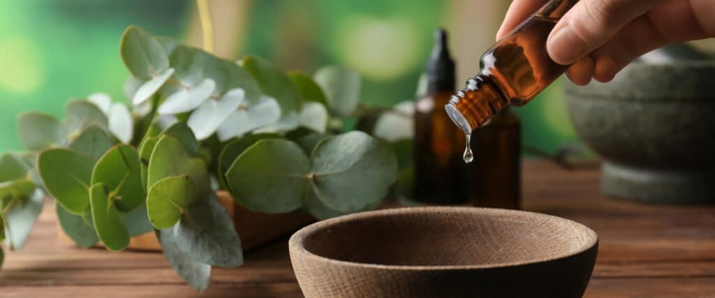 Une main verse de l'huile essentielle d'une bouteille ambrée dans un bol en bois. Feuilles d'eucalyptus et mortier en arrière-plan.
